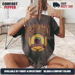 May include: A charcoal grey short-sleeve t-shirt featuring a vintage-style graphic with the text WASHINGTON FOOTBALL ESTABLISHED 1932. The design includes a central football helmet icon surrounded by a yellow laurel wreath and a maroon banner.
