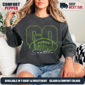 May include: A charcoal grey crewneck sweatshirt featuring a lime green graphic design. The design includes the word GO in large block letters, a football outline containing a Seattle city skyline silhouette, and the word Seattle in script below.