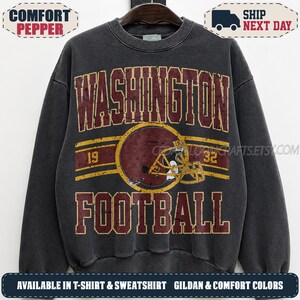 May include: A charcoal grey crewneck sweatshirt featuring a distressed graphic print. The design includes the text WASHINGTON FOOTBALL in maroon, a central maroon football helmet illustration, and the numbers 19 and 32 in a yellow horizontal band.