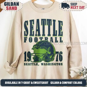 May include: A sand-colored crewneck sweatshirt featuring dark blue and lime green graphic text that reads SEATTLE FOOTBALL 1976 SEATTLE, WASHINGTON. The design includes a central illustration of a football helmet in dark blue and lime green.