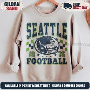May include: A sand-colored crewneck sweatshirt featuring a distressed graphic print. The design includes the text SEATTLE FOOTBALL, the numbers 19 and 76, a football helmet illustration, and a checkered pattern in navy blue and lime green.