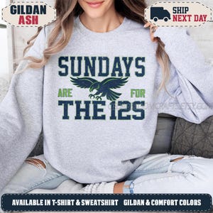 May include: A light grey crewneck sweatshirt featuring a dark blue and green graphic print. The text reads SUNDAYS ARE FOR THE 12S, centered around a stylized eagle illustration. The design has a distressed, vintage-inspired texture throughout.