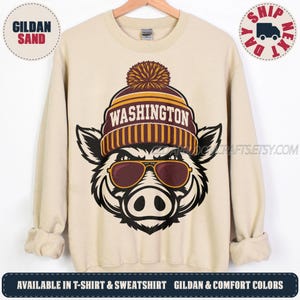 May include: A sand-colored crewneck sweatshirt featuring a graphic of a boar wearing a maroon and gold pom-pom beanie with the word WASHINGTON printed on it. The boar is also wearing brown-tinted sunglasses, rendered in a bold, black-and-white illustration style.