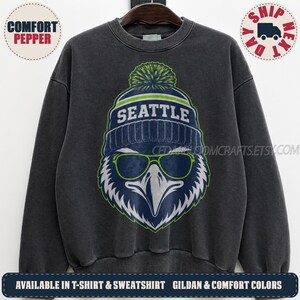 May include: A charcoal grey crewneck sweatshirt featuring a graphic of an eagle wearing a blue and green pom-pom beanie with the word SEATTLE printed on it. The eagle is also wearing lime green rimmed sunglasses, centred on the chest.