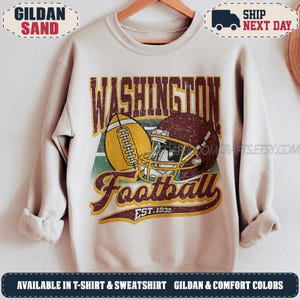 May include: A sand-coloured crewneck sweatshirt featuring a graphic print with the text WASHINGTON Football EST. 1932. The design includes a yellow football and a maroon football helmet centred over a green and white geometric background element.