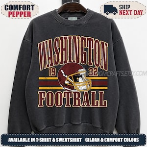 May include: A charcoal grey crewneck sweatshirt featuring a distressed graphic print. The design includes the text WASHINGTON 19 32 FOOTBALL in burgundy and yellow, centred around a burgundy football helmet with a yellow face mask.