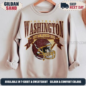 May include: A sand-coloured crewneck sweatshirt featuring a distressed graphic print. The design includes the text WASHINGTON FOOTBALL ESTABLISHED 1932 in dark red, centred above a detailed illustration of a dark red football helmet with a gold facemask.