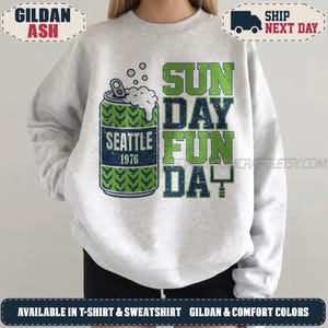 May include: A light grey crewneck sweatshirt featuring a graphic of a beer can with green and blue patterns, white foam, and the text SEATTLE 1976. To the right, the text SUNDAY FUN DAY is printed in large, distressed green and blue block letters.