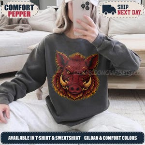 May include: A charcoal grey crewneck sweatshirt featuring a large, detailed graphic of a wild boar head with prominent tusks, rendered in shades of red, orange, and yellow. The design is centred on the chest of the long-sleeved pullover garment.