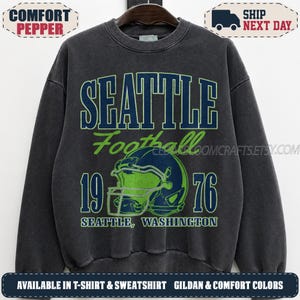 May include: A charcoal gray crewneck sweatshirt featuring a graphic print with the text SEATTLE Football 1976 SEATTLE, WASHINGTON and an illustration of a football helmet in navy blue and lime green colors on the front center of the garment.