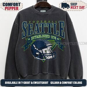 May include: A charcoal grey crewneck sweatshirt featuring a distressed graphic print. The design includes the text SEATTLE FOOTBALL ESTABLISHED 1976 in blue and green, centred above a dark blue football helmet illustration with a white face mask.