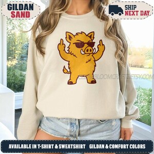 May include: A sand-coloured crewneck sweatshirt featuring a graphic of a yellow wild boar wearing dark sunglasses and giving the middle finger with both hands. The boar has a distressed, vintage-style print in dark red and yellow tones.