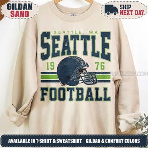 May include: A sand-colored crewneck sweatshirt featuring a graphic print with the text SEATTLE, WA, SEATTLE, and FOOTBALL in dark blue. The design includes a dark blue football helmet, the numbers 19 and 76, and horizontal green stripes.