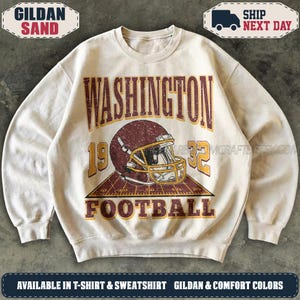 May include: A sand-coloured crewneck sweatshirt featuring a distressed graphic of a maroon football helmet on a gridiron field. The text reads WASHINGTON 1932 FOOTBALL in bold, weathered maroon and gold lettering across the front of the garment.