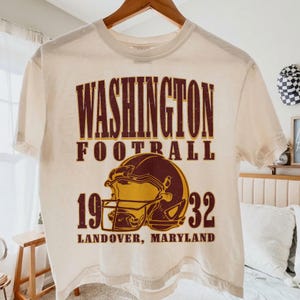 May include: A cream-coloured short-sleeve t-shirt featuring a distressed graphic in maroon and gold. The text reads WASHINGTON FOOTBALL 1932 LANDOVER, MARYLAND, centred above and below a detailed illustration of a football helmet in profile.