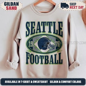 May include: A sand-coloured crewneck sweatshirt featuring a graphic design with the text SEATTLE FOOTBALL in dark blue and green. The centre displays a dark blue football helmet inside an oval frame with the numbers 19 and 76 on either side.