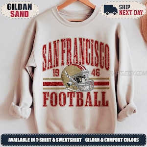 Könnte beinhalten: Ein sandfarbenes Sweatshirt mit Rundhalsausschnitt und einem abgenutzten roten und goldenen Aufdruck. Das Design zeigt den Text SAN FRANCISCO 1946 FOOTBALL sowie eine zentrale Abbildung eines goldenen Footballhelms mit rotem Streifen und weißem Gitter.