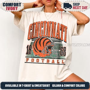 May include: An ivory short-sleeved t-shirt featuring a distressed graphic print with the text CINCINNATI 1968 FOOTBALL. The design includes an orange and black striped football helmet centred above the word FOOTBALL, all in a vintage style.