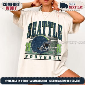 May include: An ivory t-shirt featuring a graphic print with the text SEATTLE 1976 FOOTBALL in dark blue and green. The design includes a dark blue football helmet with a face mask, set against horizontal stripes in a distressed, weathered style.
