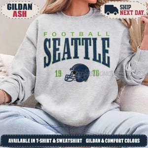 May include: A light gray crewneck sweatshirt featuring the text FOOTBALL SEATTLE in dark blue with a distressed finish. The numbers 19 and 76 are printed on either side of a dark blue football helmet graphic centered on the chest area.