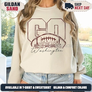 May include: A sand-coloured crewneck sweatshirt featuring a dark maroon graphic on the front. The design includes the letters GO, a football shape containing a line-art illustration of the Washington D.C. skyline, and the word Washington in script.