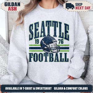May include: A light gray crewneck sweatshirt featuring a distressed dark blue and green graphic print that reads SEATTLE 1976 FOOTBALL with a central illustration of a football helmet. The design is centered on the chest of the long-sleeved garment.