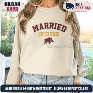 May include: A sand-colored crewneck sweatshirt featuring the maroon text "MARRIED" arched above the yellow text "INTO THIS" and a maroon graphic of a wild boar. The design is centered on the chest of the long-sleeved pullover garment.