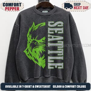 May include: A charcoal grey crewneck sweatshirt featuring a distressed graphic of a bright green eagle head on the left side and the word SEATTLE printed vertically in large, distressed grey block letters on the right side of the chest.