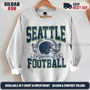May include: A light grey crewneck sweatshirt featuring a dark blue and green graphic print. The text reads SEATTLE FOOTBALL with a central illustration of a football helmet, the words SEATTLE WASHINGTON, and ESTABLISHED 1976 on a banner.