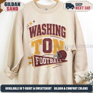 May include: A sand-colored crewneck sweatshirt featuring a graphic print with the text "WASHING TON FOOTBALL" in maroon and gold. The design includes a maroon football helmet and a pennant flag graphic with star accents in a distressed style.