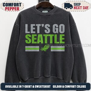 May include: A charcoal gray crewneck sweatshirt featuring the text LET'S GO in gray and SEATTLE in lime green. Below the text is a lime green eagle graphic flanked by two horizontal stripes, one lime green and one gray, on each side of the bird.