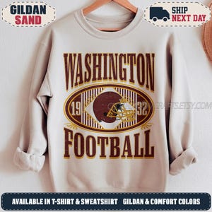 May include: A sand-coloured crewneck sweatshirt featuring a graphic with the text WASHINGTON FOOTBALL in dark red. The design includes a central oval with vertical stripes, a brown football helmet, and the numbers 19 and 32 on either side.