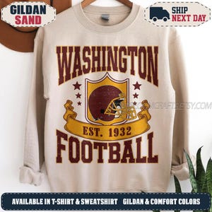 May include: A sand-coloured crewneck sweatshirt featuring a graphic print with the text WASHINGTON FOOTBALL EST. 1932. The design includes a maroon football helmet inside a shield emblem, flanked by stars, with a gold banner across the centre.