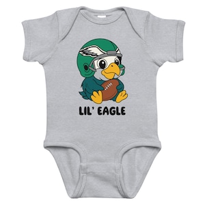 Pode incluir: Um body de bebê cinza com uma águia de desenho animado usando um capacete de futebol americano verde e segurando uma bola de futebol marrom. O texto "LIL' EAGLE" é impresso abaixo. O body é para bebês.