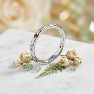 May include: A silver-toned metal band ring featuring flush-set circular gemstones, including vibrant red rubies and clear white diamonds, arranged at intervals around the circumference of the polished, smooth, and rounded metal band.