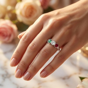 Könnte beinhalten: Ein silberfarbener Ring mit drei rund geschliffenen Steinen in einer Reihe. Der mittlere Stein ist klar und farblos, flankiert von einem leuchtend grünen Stein auf der einen und einem tiefroten Stein auf der anderen Seite, gefasst in eine polierte Metallschiene.