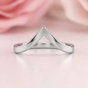 May include: A polished silver-toned chevron ring featuring a smooth, solid band that rises to a central point. The metal surface is reflective and uniform, showcasing a clean, minimalist V-shaped design that curves gently around the finger.