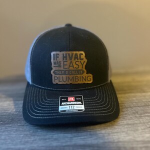 May include: A black and grey trucker hat featuring a brown leather-like patch on the front with the text IF HVAC WAS EASY THEY'D CALL IT PLUMBING. The cap includes a Richardson 112 brand sticker on the brim with white stitching details.