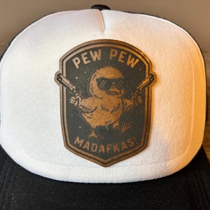 May include: A white and black trucker cap featuring a brown leather-look patch on the front. The patch displays a graphic of a chick wearing sunglasses and holding two pistols, with the text PEW PEW MADAFKAS! engraved in a dark font.