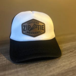 May include: A white and black baseball cap featuring a brown hexagonal patch on the front. The patch displays the text "CERTIFIED ZIP TIE MECHANIC" with a graphic of three zip ties in the centre, all rendered in a dark brown, embossed style.