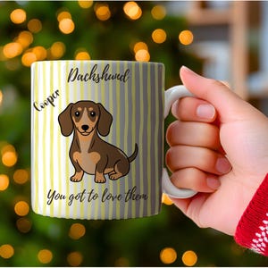 Puede incluir: Una taza de cerámica blanca con un patrón de rayas verticales amarillas y grises. La taza muestra una ilustración de un perro salchicha marrón, con el texto "Dachshund", "Cooper" y "You got to love them" impreso en letra negra.