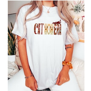 Puede incluir: Una camiseta blanca de manga corta con el texto CAT MOTHER en una fuente estilizada. Las letras están rellenas con varios patrones, incluyendo diseños florales, de cuadros y marrones sólidos. Una silueta de gato marrón se sitúa sobre el texto.