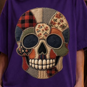 Peut inclure: Un t-shirt violet arborant un grand motif central représentant un crâne humain composé de divers motifs de tissus, notamment du tartan rouge et noir, du pied-de-poule et des imprimés floraux. Le crâne présente des orbites sombres.