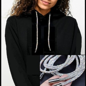May include: A black hooded sweatshirt featuring long, flexible drawstrings covered in small, reflective silver crystals. The drawstrings hang down from the hood and terminate in smooth, cylindrical silver-toned metal aglets at the ends.