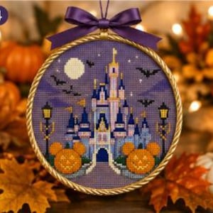 Könnte beinhalten: Eine kreisförmige Kreuzstichstickerei mit einem Schloss mit blauen und violetten Türmen, einem Vollmond und fliegenden Fledermäusen. Zwei orangefarbene Kürbisse in Form von Mausohren befinden sich an der Basis, gerahmt in einem goldenen Ring.