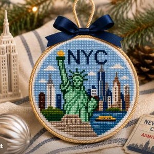 以下が含まれることがあります： 自由の女神像とニューヨーク市のスカイライン、そしてダークブルーの「NYC」の文字が刺繍された円形のクロスステッチオーナメント。刺繍はツイストロープの縁取りが施された木製の枠に収められ、ダークブルーのサテンリボンが飾られています。