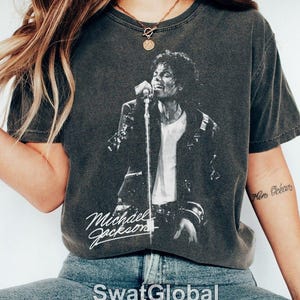 May include: A charcoal gray short-sleeve t-shirt featuring a black and white graphic print of Michael Jackson singing into a microphone. The shirt has a vintage-style washed finish and includes the name Michael Jackson in white script text.