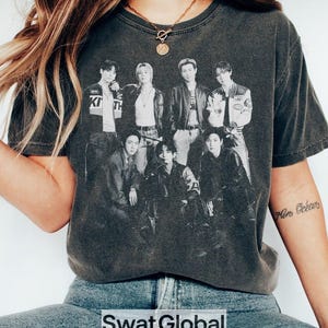 Pode incluir: Uma t-shirt de manga curta em cinzento-carv&atilde;o com uma impress&atilde;o gr&aacute;fica a preto e branco de um grupo de sete pessoas a posar juntas. A t-shirt tem um decote redondo e um aspeto vintage lavado, exibindo um design fotogr&aacute;fico detalhado.