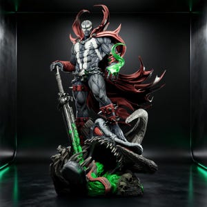 May include: A detailed Spawn character statue featuring a muscular figure in a grey suit with white chest markings, a flowing red cape, and spiked red gauntlets. The figure holds a large sword and stands atop a dark, textured base with green accents.