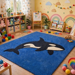 Puede incluir: Una alfombra rectangular azul que presenta un gráfico grande y centrado de una orca en blanco y negro. La alfombra tiene una superficie texturizada y afelpada, y está colocada en el centro de una habitación llena de diversos juguetes infantiles.